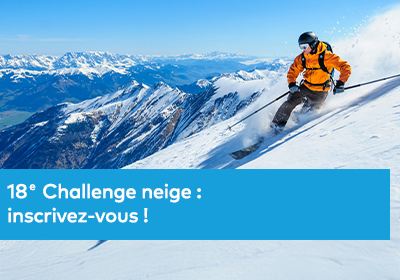 Photographie d’un skieur équipé d’une combinaison orange descendant une pente enneigée en montagne, avec un panorama de sommets alpins sous un ciel bleu.